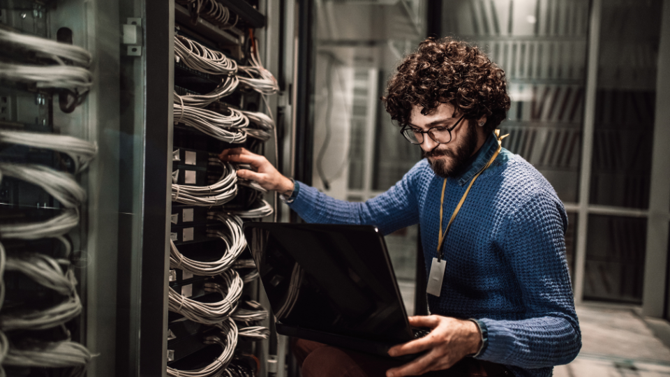 IT technician with curly hair and glasses checks server racks in a data center, laptop in hand.